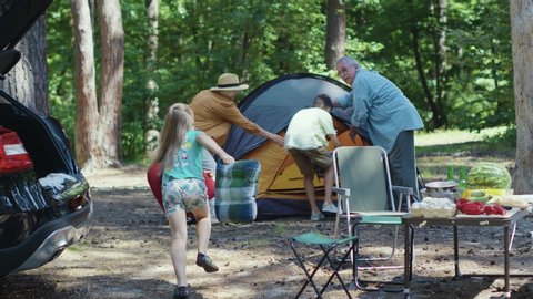 Couples of grandparents and kids putting up camptent in forest. Helpful children run to the tent decorateing it with pillows. Camping with family. Picnic. Summertime.