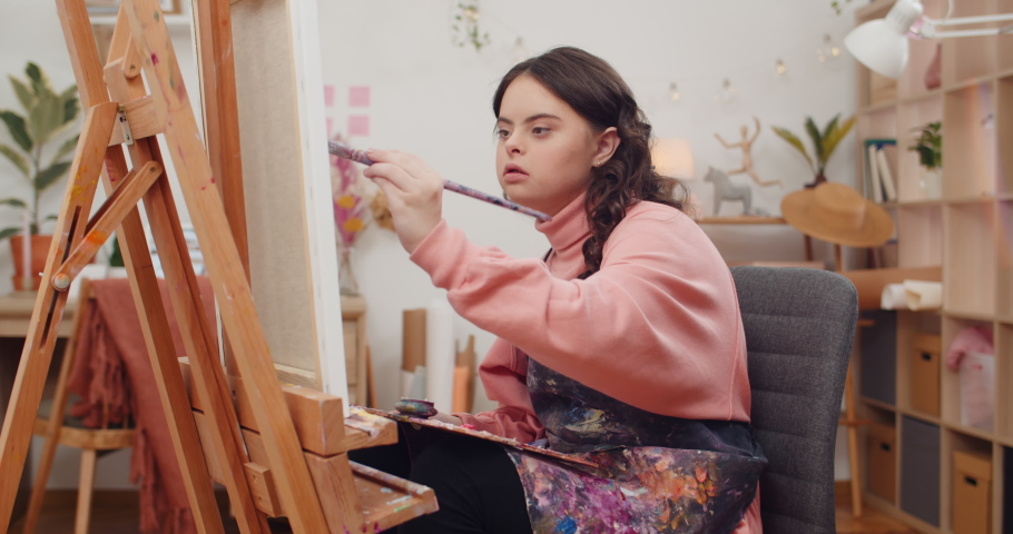 Teen wearing artists apron painting and putting brush from one hand to other at her room. Girl with down syndrome holding palette on knees while creating picture in front of molbert Royalty-Free Stock Footage #1058327317
