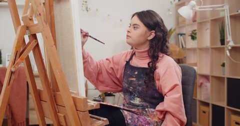 Teen wearing artists apron painting and putting brush from one hand to other at her room. Girl with down syndrome holding palette on knees while creating picture in front of molbert