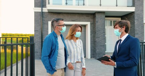 Caucasian young happy couple in medical masks buying house at suburb and talking with male real-estate agent holding tablet device in hands. Outdoor. Coronavirus pandemic concept Using gadget computer