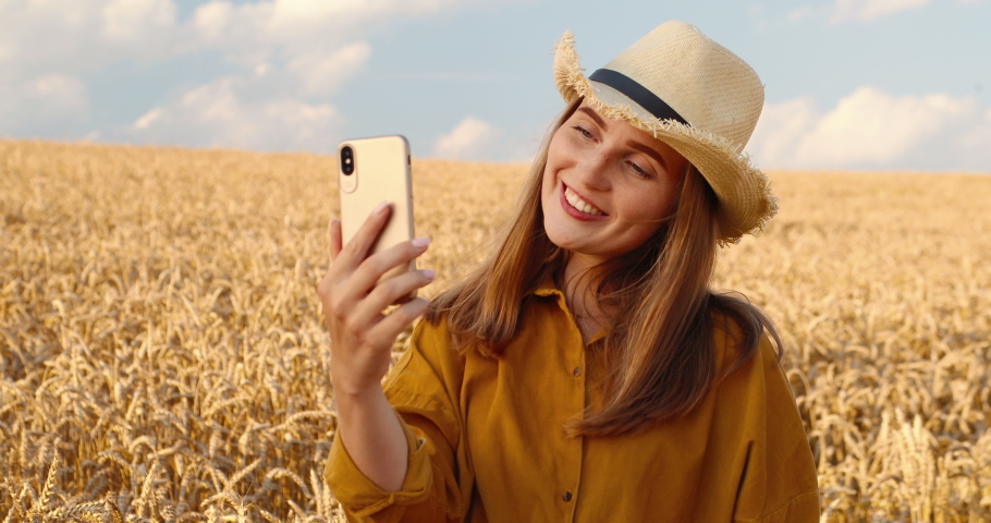 Beautiful Caucasian happy girl in hat having video call on smartphone while standing in wheat field in village. Pretty young woman videochatting on cellphone outdoors in rural area. Video chat concept