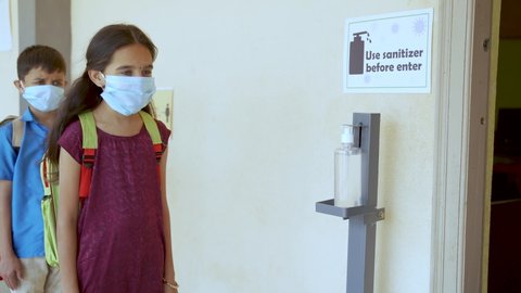 Kids with medical mask using hand sanitizer before entering classroom while maintaining social distancing - concept of back to school or school reopen and coronavirus or covid-19 safety measures.