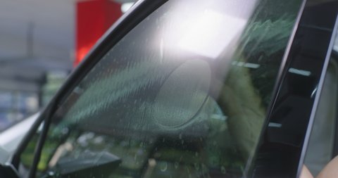 Bearded man spraying water on tinted window of car and cleaning glass while working in garage
