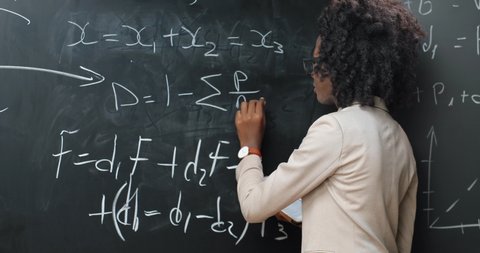 Back view on African American young woman teacher at school writing formulas and mathematics laws on blackboard. School concept. Female lecturer in glasses explaining physics laws. Rear.