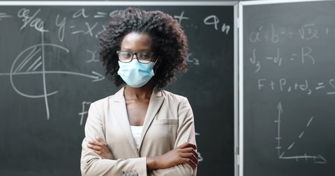 Portrait of young African American female teacher in glasses and medical mask looking at camera and crossing hands in classroom. Blackboard with math formulas on background. Pandemic schooling.