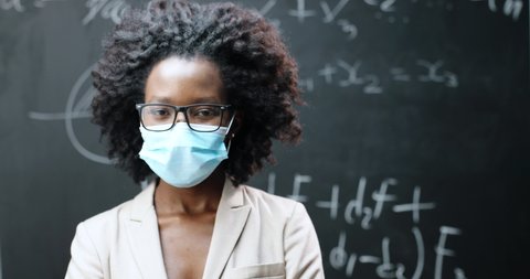 Portrait of young African American female teacher in glasses and medical mask looking at camera in classrom. Blackboard with formulas on background. Coronavirus concept. Pandemic schooling.