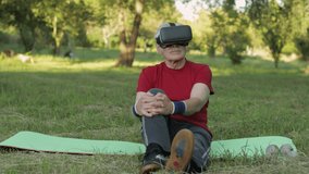 Elderly man in VR headset helmet sitting on sport mat outdoors in park. Online sport workout for senior grandparents people. Old grandfather watching virtual reality 3D 360 video game