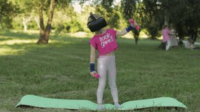 Athletic child in VR headset helmet making fitness exercises with dumbbells outdoors in park. Online workout for kids. Little caucasian girl kid watching virtual reality 3D 360 video game on sport mat