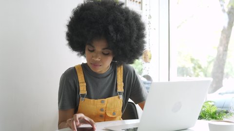 Young stylish hipster gen z African American female student with afro hair using laptop computer and smartphone sitting at table at home, in cafe, remote studying, elearning or working in internet.