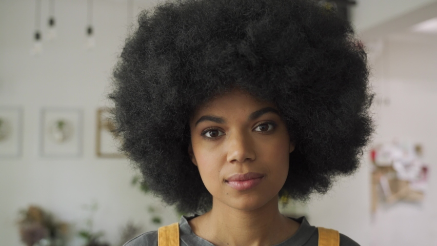 Confident stylish African American gen z hipster female student with Afro hair looks at camera, dreaming, standing in cozy cafe interior. Proud mixed race young woman headshot portrait. | Shutterstock HD Video #1058339590