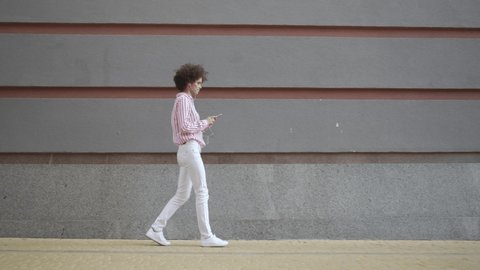 Smiling young european woman walking on urban city street against grey wall holding smart phone. Happy millennial curly lady wearing earphones using mobile apps listening music on cellphone outdoor.