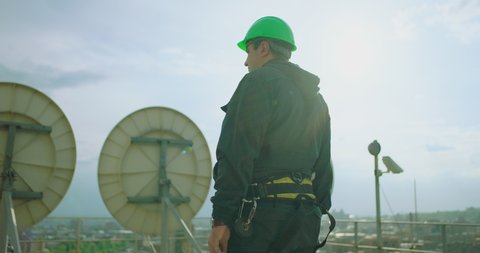 Side view of portrait of engineer . Professional telecommunication industry engineer looking horizon of city. Worker wearing safety uniform and hard hat on unfocused background with city and antennas
