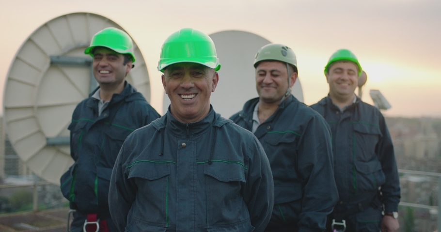 Portrait of group of professional telecommunication industry engineers smiles and looks at the camera . Workers wearing safety uniform and hard hat on unfocused background with sky and antennas . Royalty-Free Stock Footage #1058342944