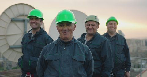 Portrait of group of professional telecommunication industry engineers smiles and looks at the camera . Workers wearing safety uniform and hard hat on unfocused background with sky and antennas .