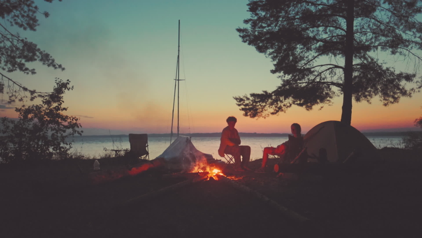 Couple, sailing boat and bonfire. Young couple sit near the bonfire with sailing boat beached on the background