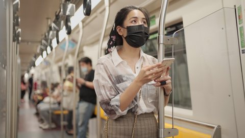 Asian woman in black protective mask using smartphone, stand leaning on the pole, covid-19 new normal, woman in subway metro, self protection on public transport, social distancing, lost wondering 