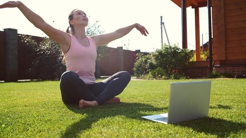 Online meditation lesson. Young woman in sportswear in lotus position looking at laptop screen and gives online yoga lessons, meditating outdoors in the morning on a sunny day.