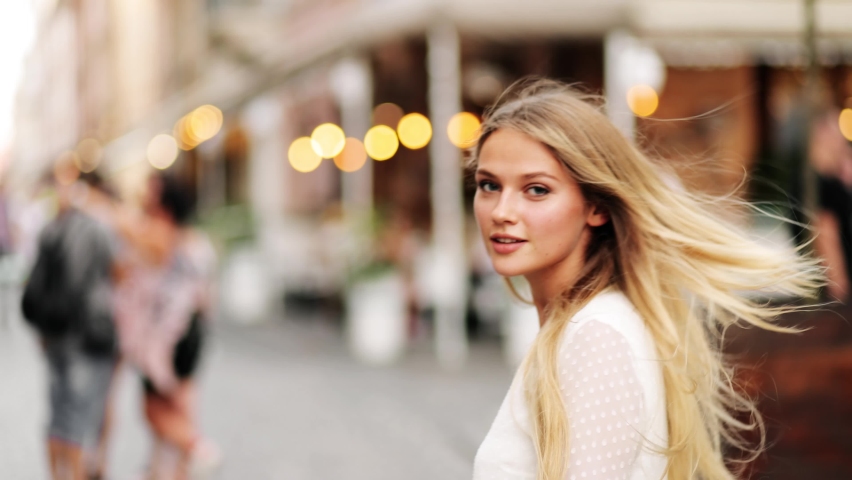 Back view of seductive hot blonde woman walking on city street in slow motion, turning and smiling straight to camera