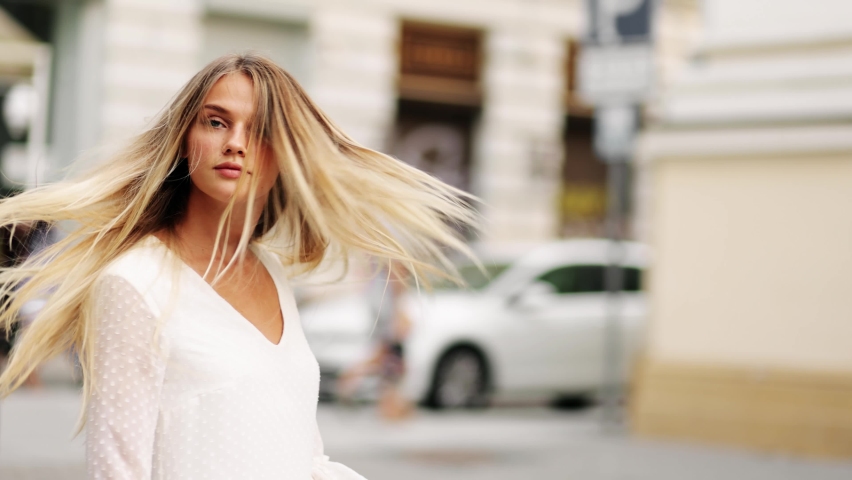 Young beautiful blonde woman in a white dress spinning in the center of a city. Attractive fashion model with flying hair. Beautiful girl woman shakes hair. Slow motion