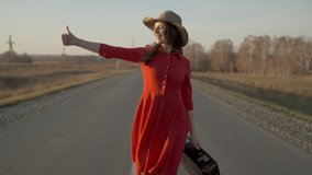 Young happy beautiful traveler tourist woman in red dress and hat with suitcase at outdoors hitchhiking in summer rural road. Travel hitchhiker tourism trip traveling nature vacation. 4 K slow-mo