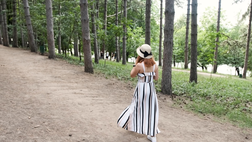Red haired young woman with a hat on her head is walking in the park while is drinking a cup of coffee and talking on phone. 
