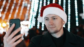 Cheerful man taking video call from friends on mobile phone, dressed in christmas hat. Christmas tree, people and lights on background