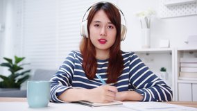 Asian Female College Student looking at camera watching online lesson and taking notes while studying online. Teenager video chat and e-learning with teacher. Distance Education Concept