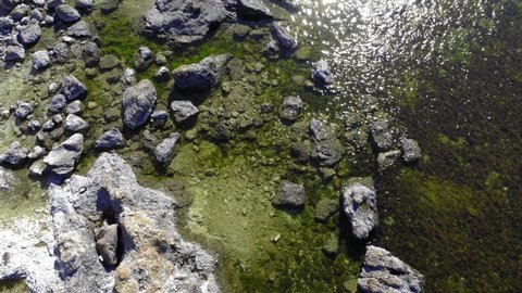 Aerial view over stones in shallow water, sun glittering on the water surface, on a warm, summer day, in Faro, Gotland, Sweden - low, tilt up,, drone shot