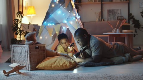 African American father holding flashlight and reading fairy tale to cute little son before going to bed while he lying on the floor under teepee tent decorated with lights in dark kids room