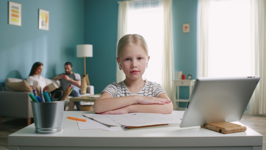 Sad girl portrait, sitting at work desk at home, looking at camera, serious and focused, in wireless headset, parents are on the background, online education concept, Slow motion.