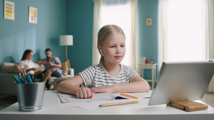 Small happy girl is sitting at work desk at home, has online lesson, raising hand to answer, use tablet computer, gadget, parents are busy with their affairs, Foreground, Slow motion.