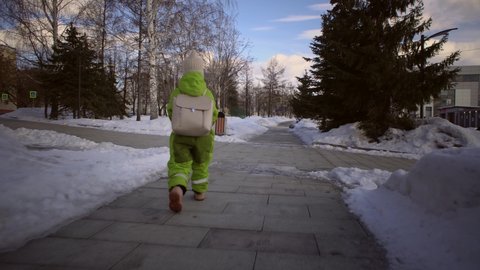 View from the back of an elementary school pupil in green jumpsuit walks through snow-covered park to school on clear day.