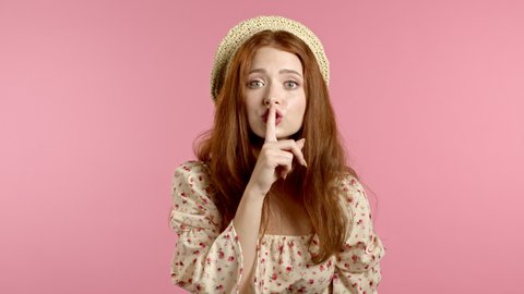 Beautiful woman in floral dress holding finger on her lips over pink background. Gesture of shhh, secret, silence. Close up.