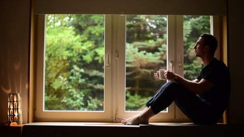 Стоковое видео: Man drinking tea from steaming cup sitting on windowsill. Man on windowsill near large window, nice interior is visible.