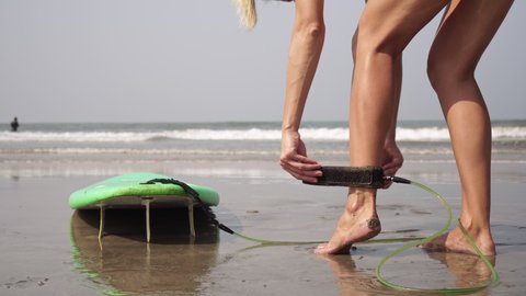 A young woman puts a surfboard leash on her leg before entering the sea