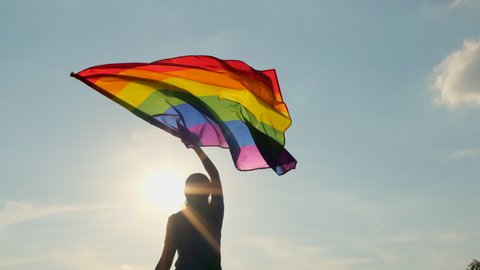 Rainbow LGBT flag on sunset blue sky. Female woman holding lgbt gay pride flag in hands -lesbian, gay, bisexual, transgender social movements. Concept of happiness freedom love same-sex couple, 4 K