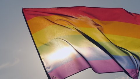 Rainbow LGBT flag on sun blue sky close-up. Female holding lgbt gay pride flag in hands -lesbian, gay, bisexual, transgender social movements, Concept happiness freedom love same-sex couple 4 K slowmo