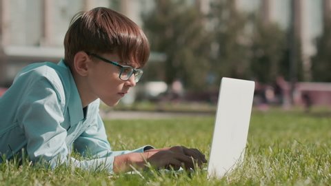 Online education, Back to school, Happy schoolboy, Learn lessons. Schoolboy doing homework using laptop while lying on grass near school