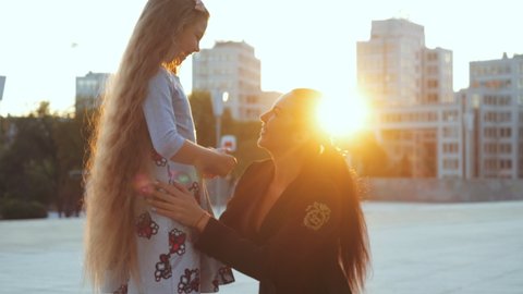 Kharkov, Ukraine - 08/22/2020: Back view little girl with long blond hair runs to meeting with young woman in classic suit, hugs her mother, rejoices at return of her older sister, sunset background