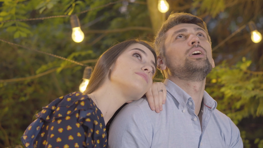 Close-up portrait of happy man and woman looking up and talking. Romantic Caucasian partners dating outdoors in summer evening. Garland lights shining at the background.