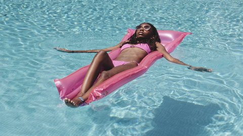 Full body focused shot of a black woman in slender body as she lies down on a pink inflatable on the pool