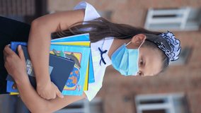 vertical video. outdoors portrait of schoolgirl, in medical protective mask, with books in her hands, on background of school. Back to school after covid19 outbreak.