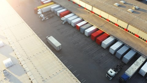 Semi truck with cargo trailer is travelling on a parking lot along a warehouse of a logistics park.  Aerial following shot 