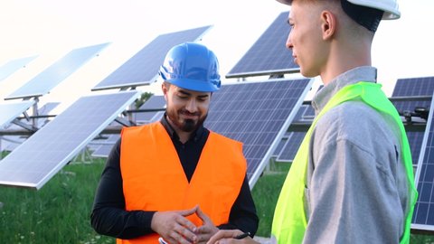 The portrait of two engineers with the project plan at the solar farm, in special uniform, discusses the installation of sunny African-American electricity batteries of slow movement of the