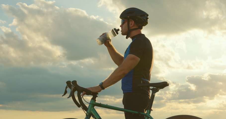 The cyclist is standing on the edge of the road and drinking water from a bottle Sunset in the background. Close-up shooting. 4K Royalty-Free Stock Footage #1058417251