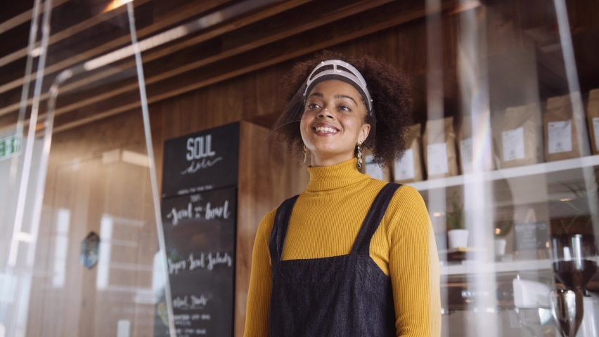Low Medium Close up, Female African coffee shop owner wearing face visor welcoming a customer Royalty-Free Stock Footage #1058419360