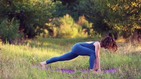 Sporty young woman doing yoga exercise on mat outdoor.Beautiful white girl in 20s wearing blue body suit working out,stretching in green park at sunset.Healthy lifestyle & female beauty concept