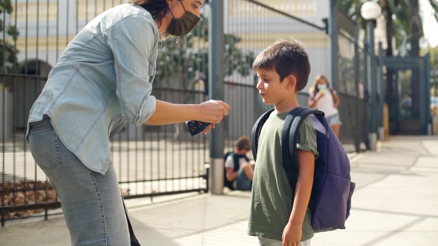 Caucasian woman straightens the protective mask on her son. They kiss on their masks and greet each other. Elementary student's first day of school during coronavirus pandemic. Concept back to school. | Shutterstock HD Video #1058428777