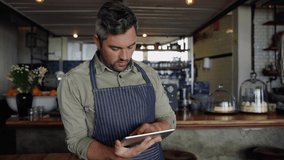 Clip of handsome cafe owner scrolling on tablet while leaning on counter of trendy coffee shop.