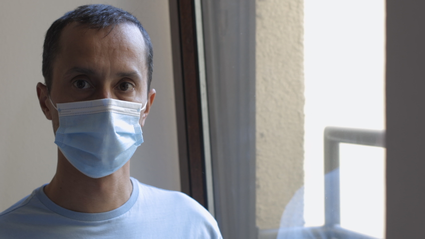 Close-up of an adult man wearing a face mask at home, looking out the window. Indoor scene expressing isolation, health concern, and emotional stress during the COVID-19 pandemic.